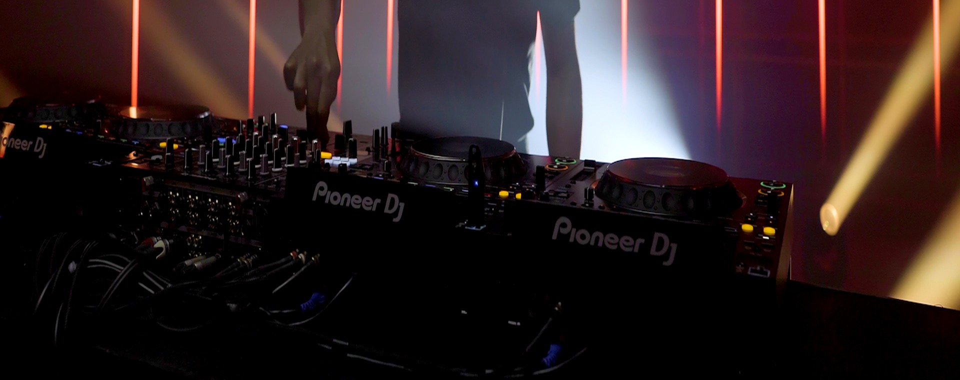 Real Pioneer DJ booth setup with CDJ players, mixer controls, stage lighting, and performance cables